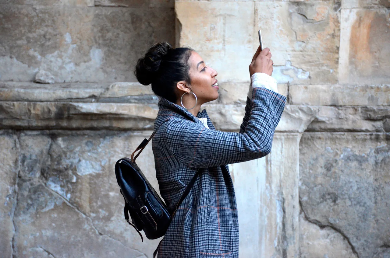 Una mujer sostiene un smartphone frente a su rostro junto a un muro de piedra.
