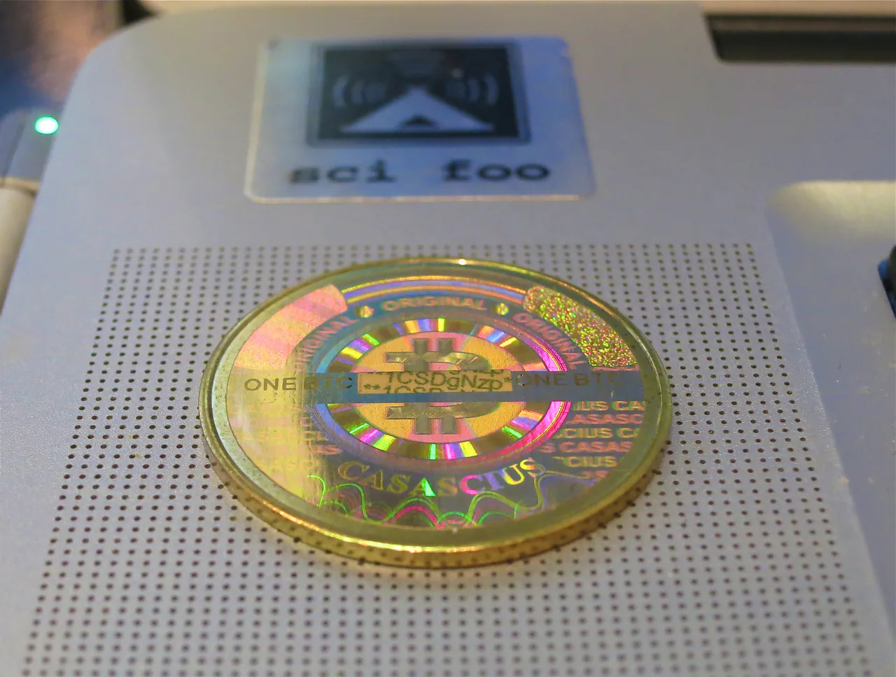 Una moneda Casascius de color dorado con una pegatina holográfica multicolor sobre un portátil.
