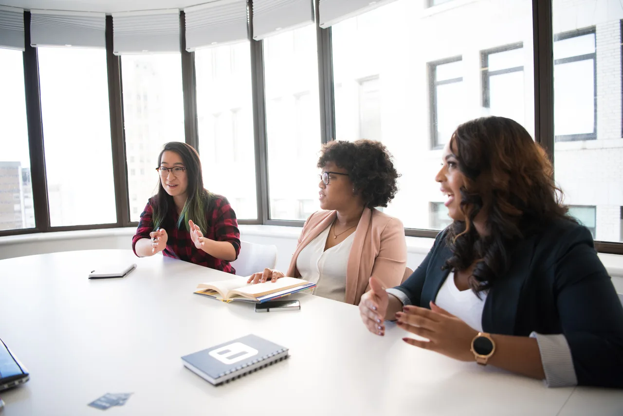 Three women talk around a work table with notebooks.