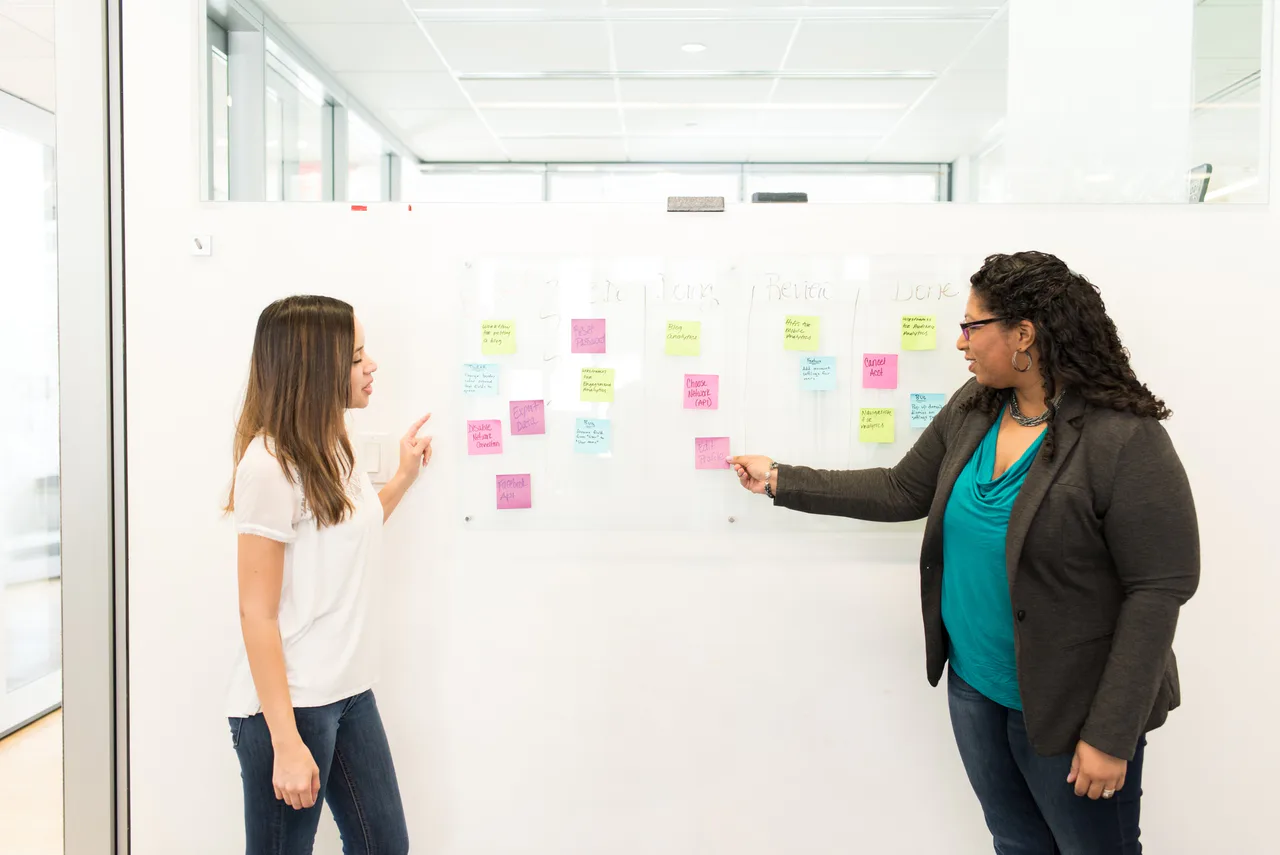 Two women discuss sticky notes on a whiteboard.