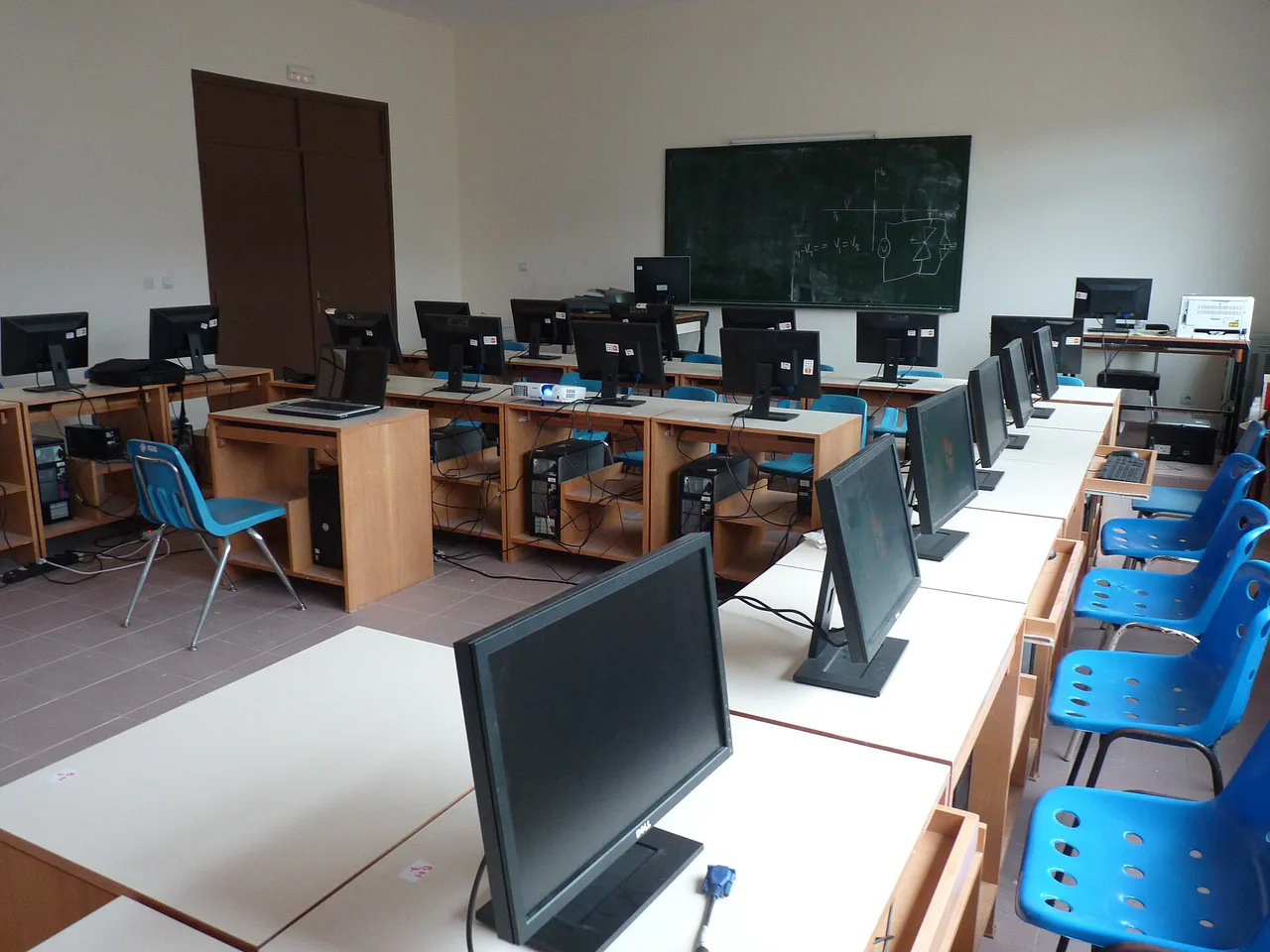 Filas de ordenadores y sillas azules en un aula informática vacía.