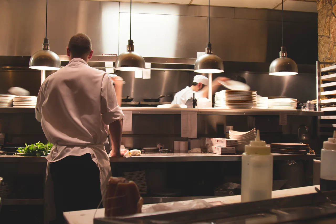 Un cocinero trabaja en una cocina profesional frente a la zona de servicio y otros compañeros