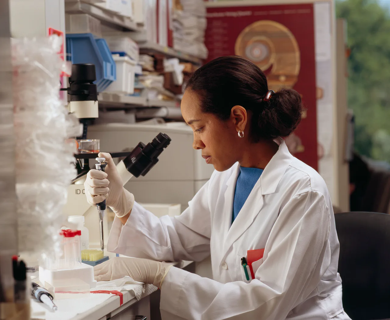 Una investigadora con bata y guantes utiliza una pipeta junto a un microscopio en un laboratorio.