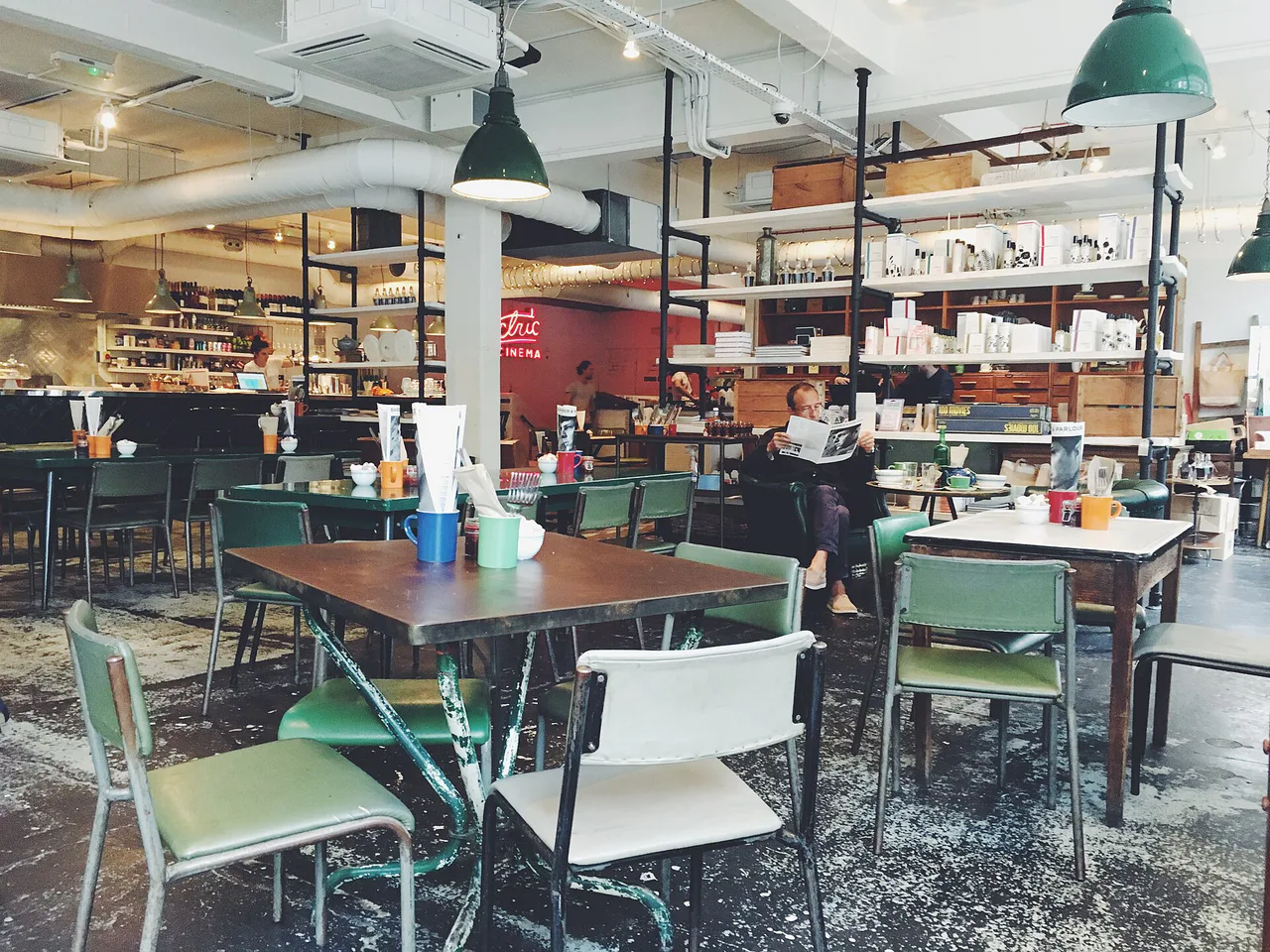 A man reads a newspaper in a café with empty tables and chairs.