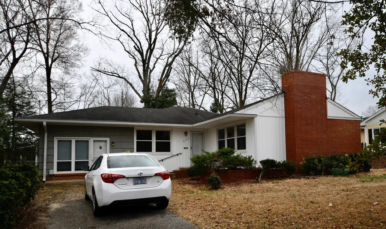 Casa de una planta con fachada clara, chimenea de ladrillo y un coche blanco aparcado delante.