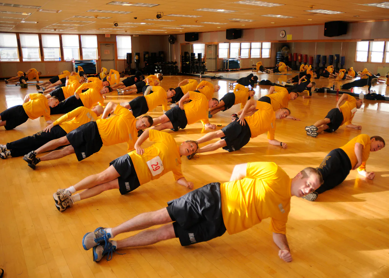Un grupo con camisetas amarillas realiza ejercicios apoyado de lado en el suelo de un gimnasio.