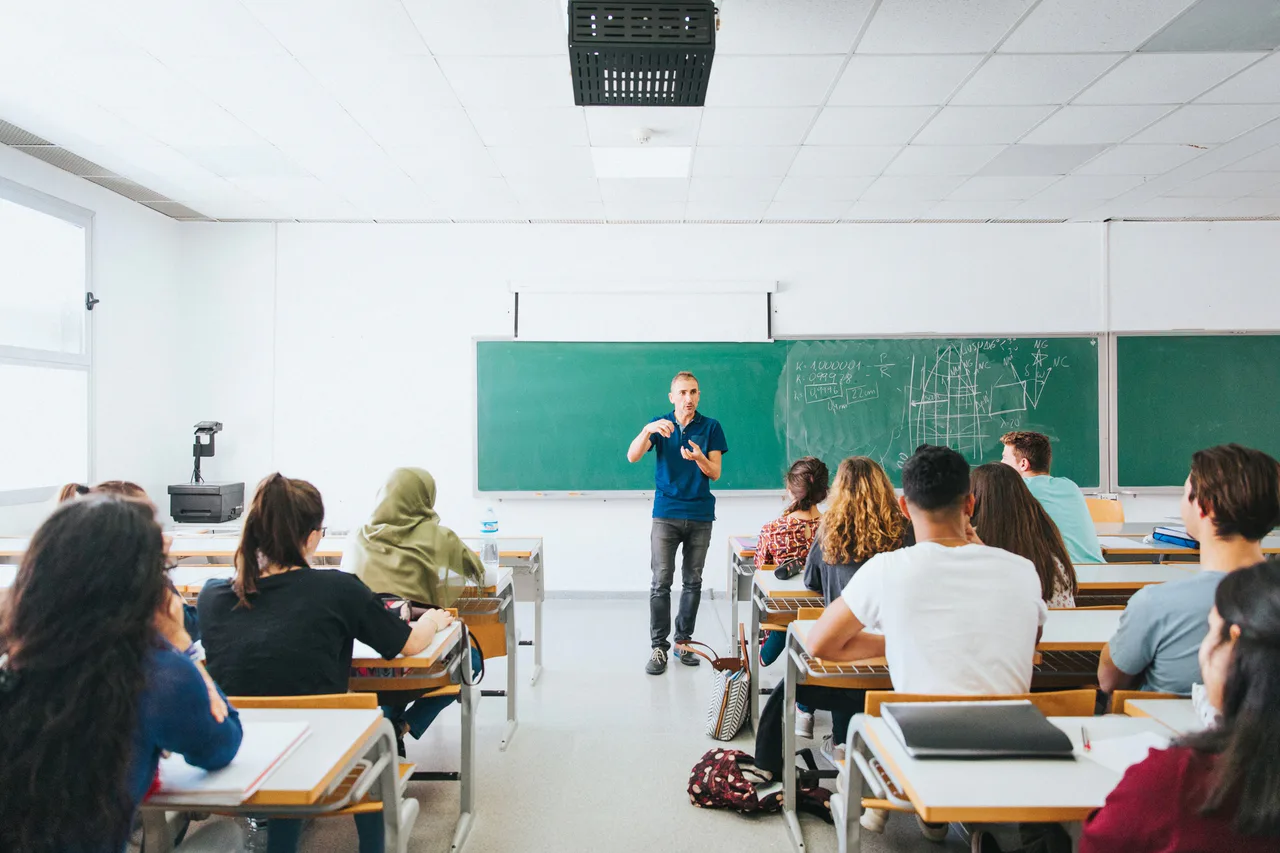 A teacher beside a chalkboard and students seated in a classroom.