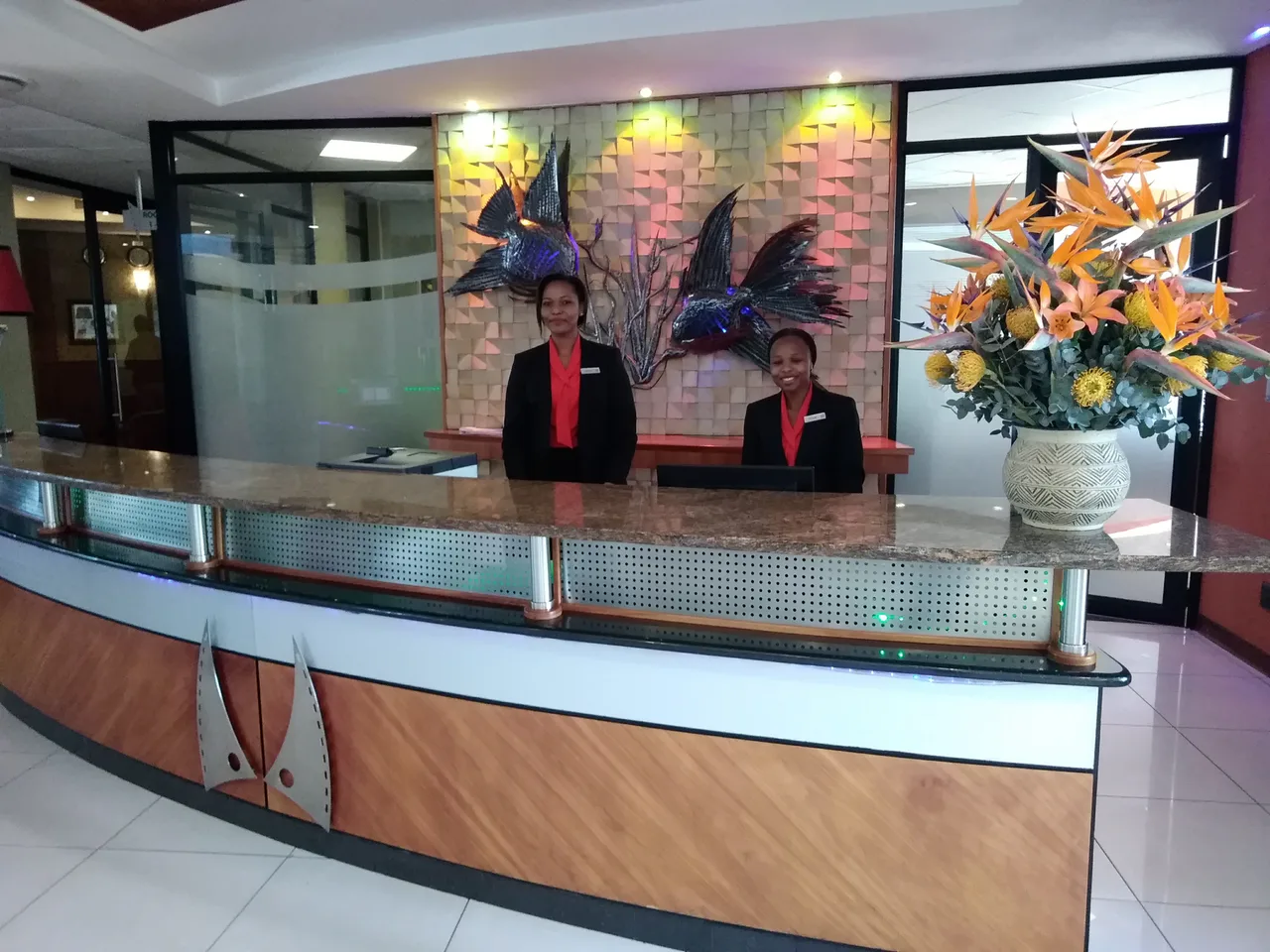 Two uniformed people behind a hotel reception desk with a computer and flowers