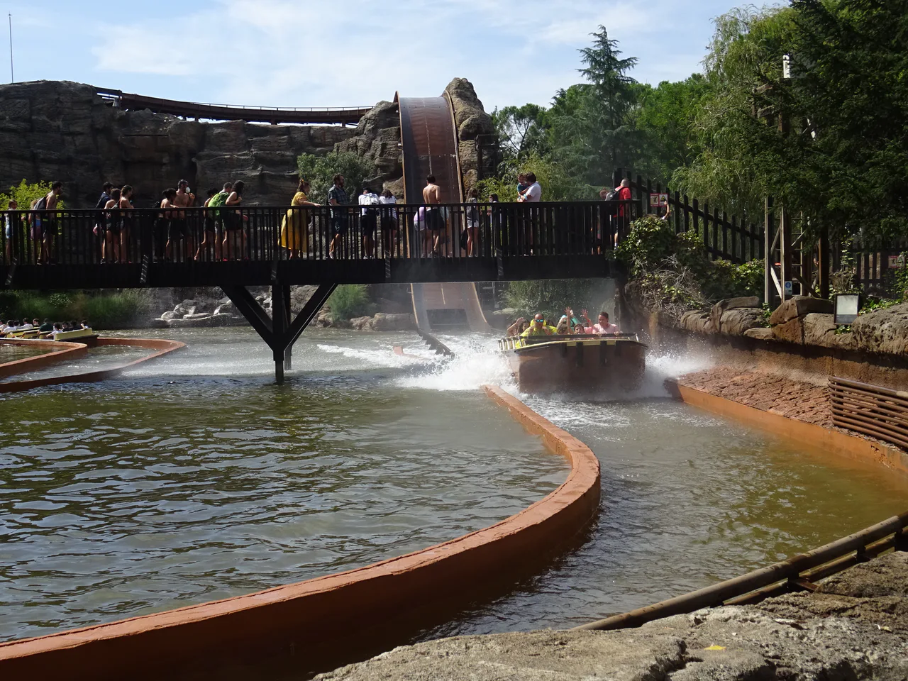 Una barca con pasajeros avanza por un canal de agua junto a un puente con personas y una rampa.
