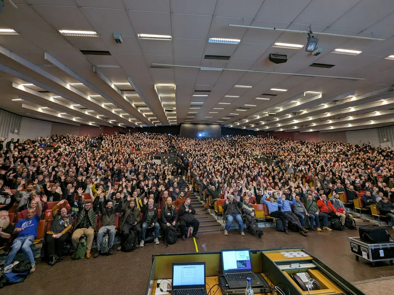 Público de FOSDEM 2024 sentado en un gran auditorio visto desde la mesa de los ponentes.