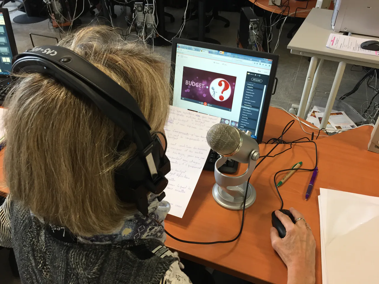 A person wearing headphones faces a desktop microphone, laptop and handwritten sheet