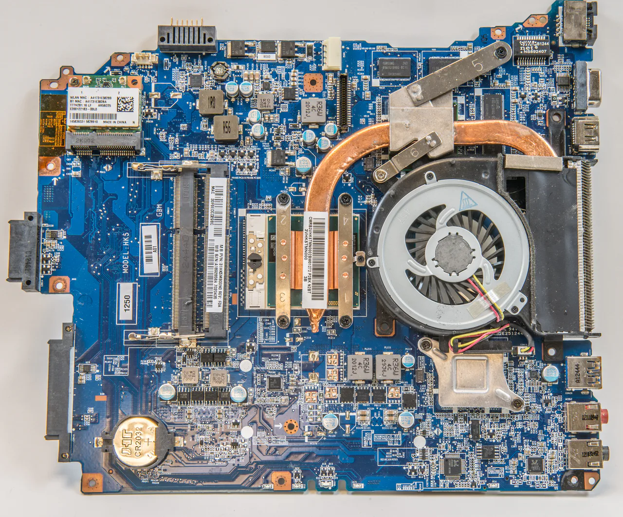 Placa base azul de un portátil con chips, ranuras de memoria, ventilador y un tubo de refrigeración de cobre.