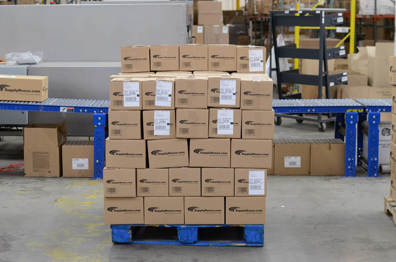 Un palé de cajas rotuladas SupplyHouse.com está junto a transportadores de rodillos en un almacén.