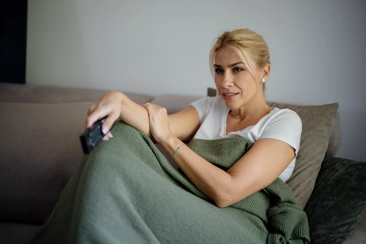A woman uses a remote control on a sofa while covered with a blanket