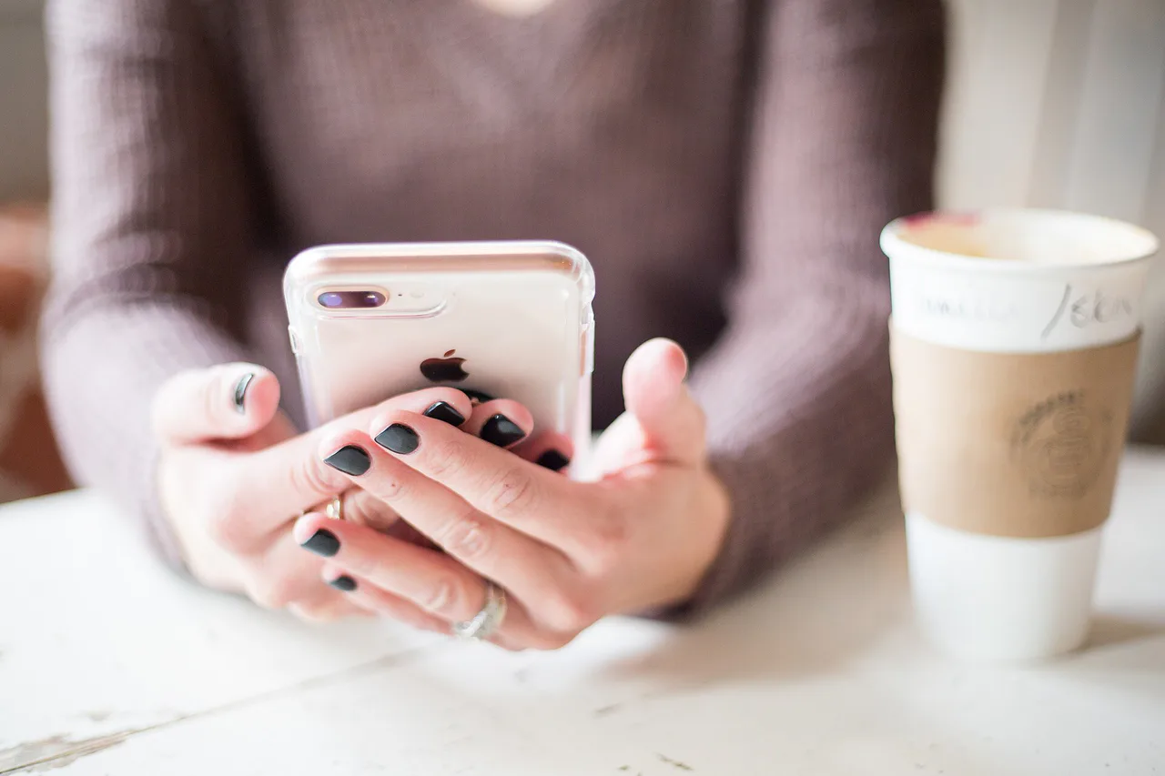 Unas manos sostienen un teléfono móvil junto a un vaso de café sobre una mesa.