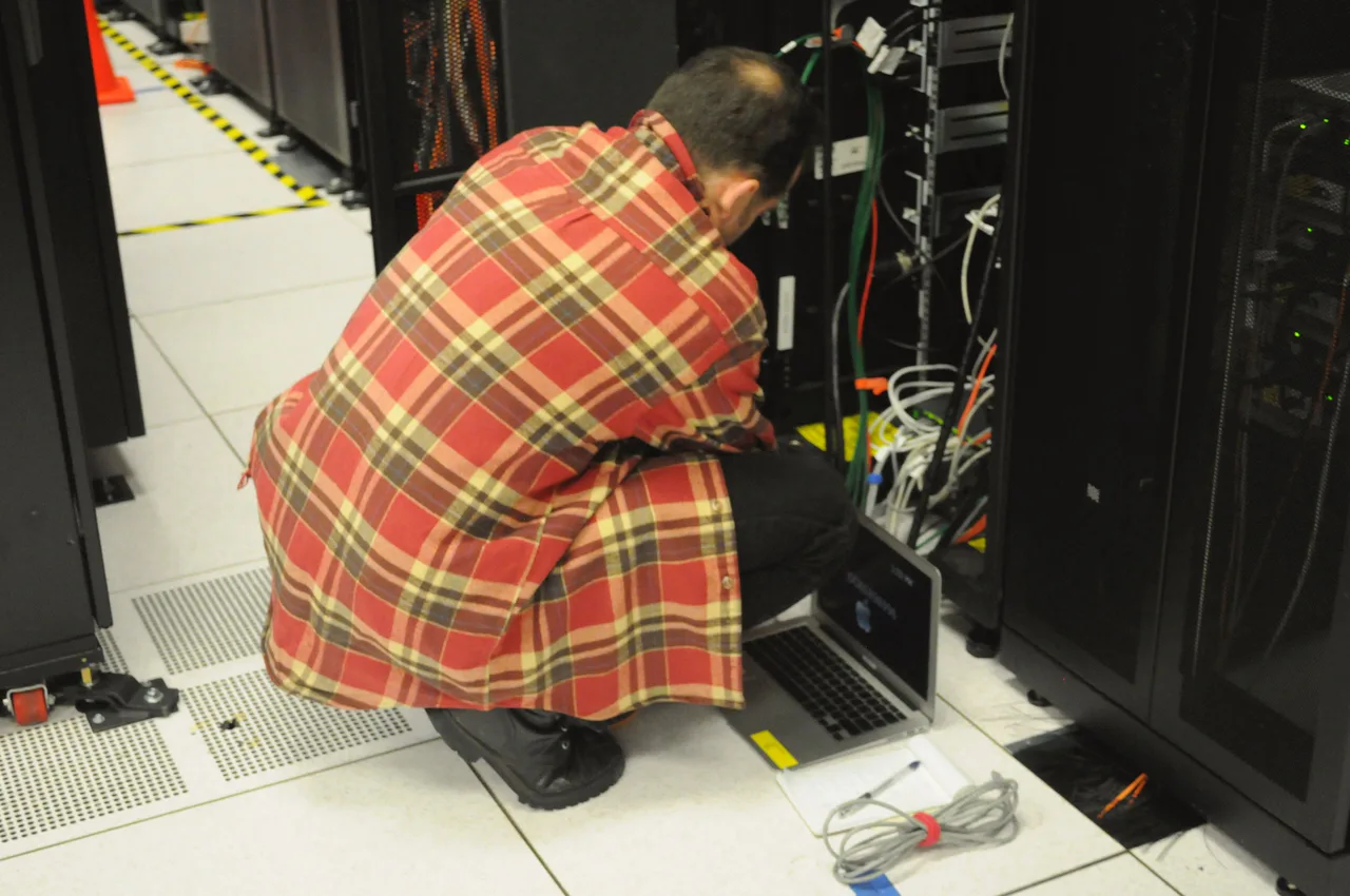 A person works with a laptop beside data centre equipment.