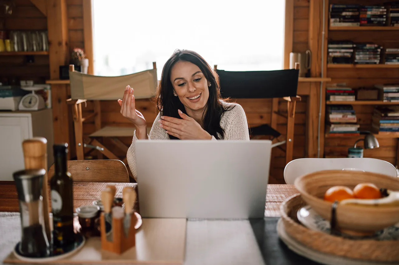A woman smiles and gestures in front of a laptop on a wooden table.